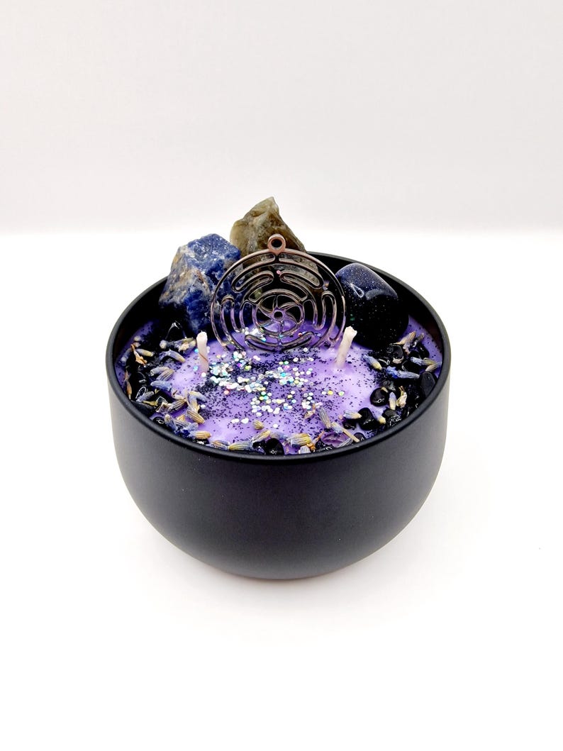Puede incluir: Un tarro de vela negro con una parte superior de cera morada. El tarro contiene cristales, purpurina y un amuleto de metal. El amuleto es un c&iacute;rculo con un dise&ntilde;o geom&eacute;trico.
