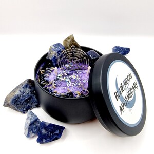 Puede incluir: Una lata de vela negra con una vela morada en su interior. La vela tiene un amuleto plateado en la parte superior y est&aacute; rodeada de piedras azules y verdes. La tapa de la lata es negra con una etiqueta blanca que dice "Blue Moon Apothecary".