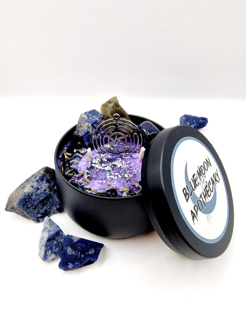 Puede incluir: Una lata de vela negra con una vela morada en su interior. La vela tiene un amuleto plateado en la parte superior y est&aacute; rodeada de piedras azules y verdes. La tapa de la lata es negra con una etiqueta blanca que dice "Blue Moon Apothecary".
