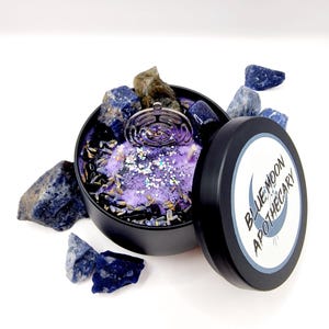 Puede incluir: Una vela morada con un amuleto plateado y cristales azules en un recipiente de hojalata negro. La tapa de la lata tiene una etiqueta blanca con el texto "BLUE MOON APOTHECARY".