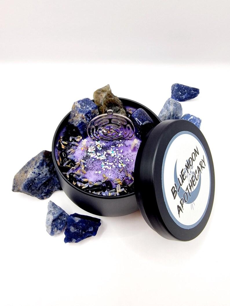 Puede incluir: Una vela morada con un amuleto plateado y cristales azules en un recipiente de hojalata negro. La tapa de la lata tiene una etiqueta blanca con el texto "BLUE MOON APOTHECARY".
