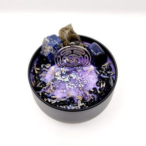 Puede incluir: Vela morada con un amuleto plateado, cristales azules y grises, y detalles negros y lavanda en una lata negra.