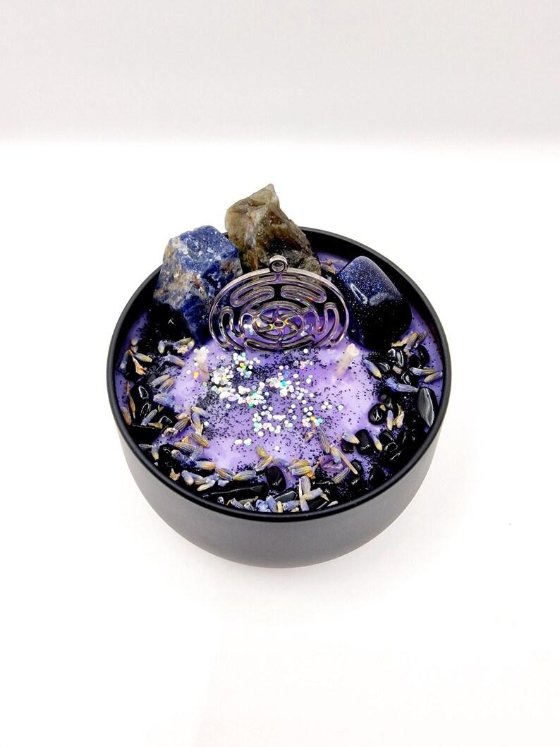 Puede incluir: Vela morada con un amuleto plateado, cristales azules y grises, y detalles negros y lavanda en una lata negra.