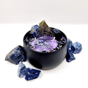 Puede incluir: Un tarro de vela negro con una vela morada y brillante en su interior. La vela tiene un dije de metal con un dise&ntilde;o geom&eacute;trico en la parte superior. El tarro est&aacute; rodeado de piedras azules y marrones.