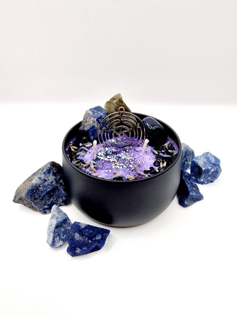Puede incluir: Un tarro de vela negro con una vela morada y brillante en su interior. La vela tiene un dije de metal con un dise&ntilde;o geom&eacute;trico en la parte superior. El tarro est&aacute; rodeado de piedras azules y marrones.