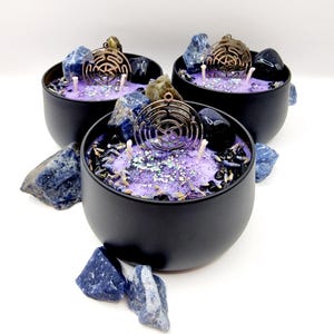 Puede incluir: Tres tarros de velas negros llenos de cera morada y un amuleto de metal de color cobre. Los tarros est&aacute;n llenos de piedras azules y negras y purpurina.