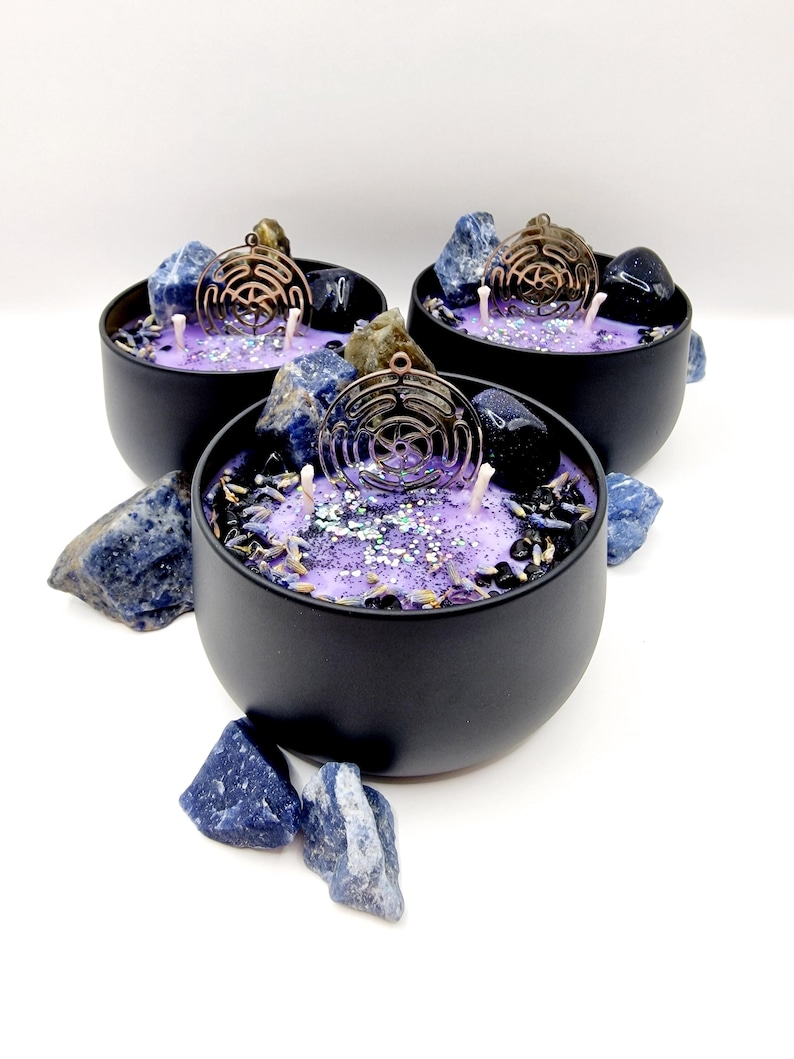 Puede incluir: Tres tarros de velas negros llenos de cera morada y un amuleto de metal de color cobre. Los tarros est&aacute;n llenos de piedras azules y negras y purpurina.