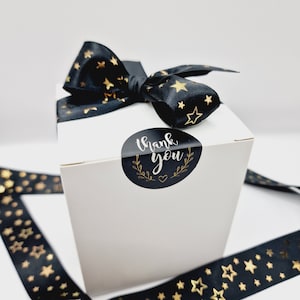 Puede incluir: Una caja de regalo blanca con una cinta negra con estrellas doradas. La caja tiene una pegatina negra y dorada que dice "Gracias".