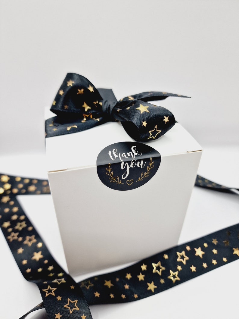 Puede incluir: Una caja de regalo blanca con una cinta negra con estrellas doradas. La caja tiene una pegatina negra y dorada que dice "Gracias".