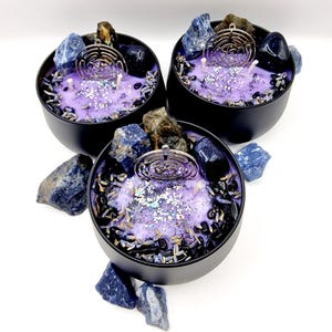 Puede incluir: Tres velas moradas y negras con cristales incrustados y un amuleto plateado en forma de laberinto. Las velas est&aacute;n en recipientes negros y est&aacute;n rodeadas de piedras azules y negras.