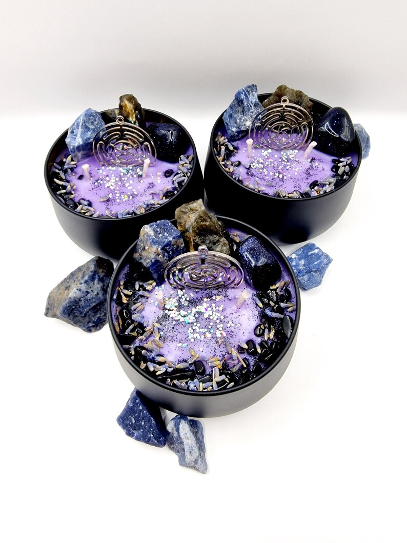 Puede incluir: Tres velas moradas y negras con cristales incrustados y un amuleto plateado en forma de laberinto. Las velas est&aacute;n en recipientes negros y est&aacute;n rodeadas de piedras azules y negras.