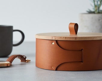 May include: A round, brown leather box with a wooden lid and a leather handle. The box has brass accents and is sitting on a gray surface.