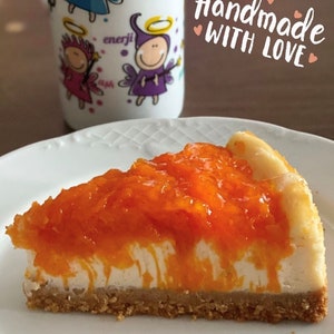 May include: A slice of cheesecake topped with a layer of orange marmalade. The cheesecake has a creamy white filling and a graham cracker crust. The text "Handmade with Love" is written in pink on a brown background.