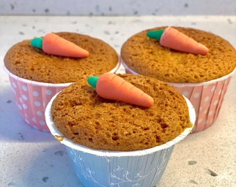 May include: Three carrot cupcakes in decorative paper cups. Each cupcake has a light brown, textured cake base and is topped with a small, orange carrot-shaped decoration with a green stem. The cupcake liners are pink with white polka dots and light blue with a floral design.