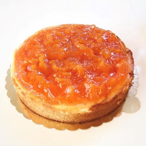 May include: A round cheesecake topped with a layer of orange marmalade. The cheesecake is on a gold paper doily.