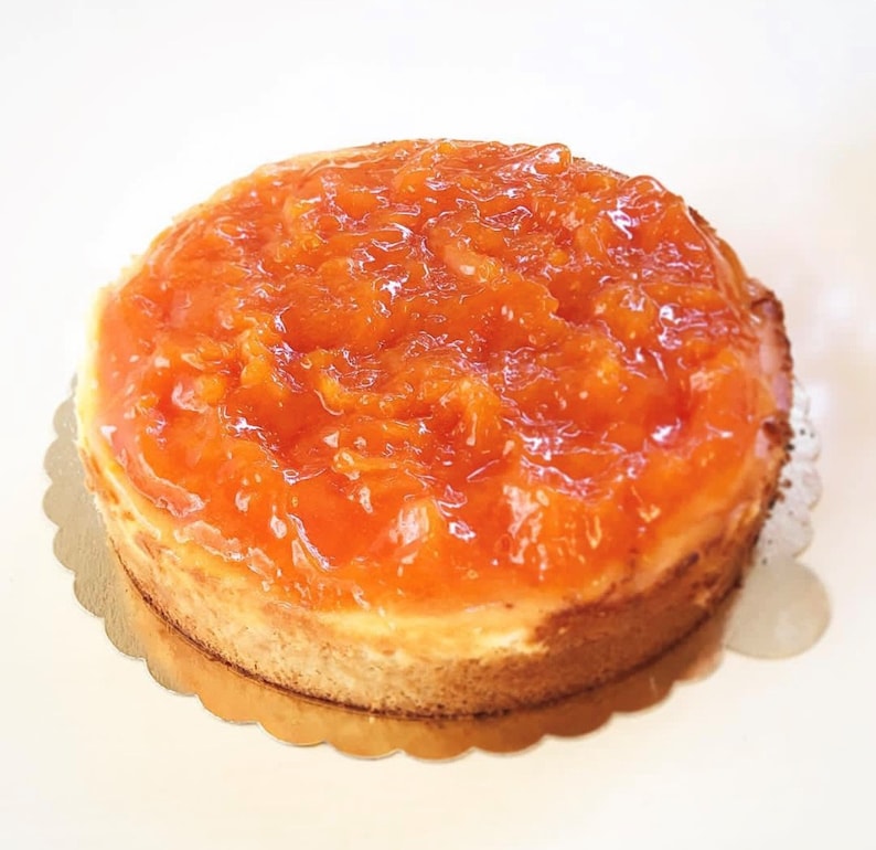 May include: A round cheesecake topped with a layer of orange marmalade. The cheesecake is on a gold paper doily.
