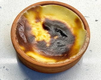 May include: A small, round, terracotta bowl filled with a creamy, yellow custard dessert. The custard has a slightly browned, caramelized top.