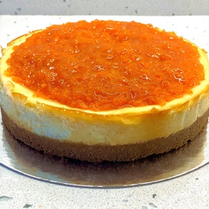 May include: A round cheesecake with a layer of orange marmalade on top. The cheesecake is on a silver plate.