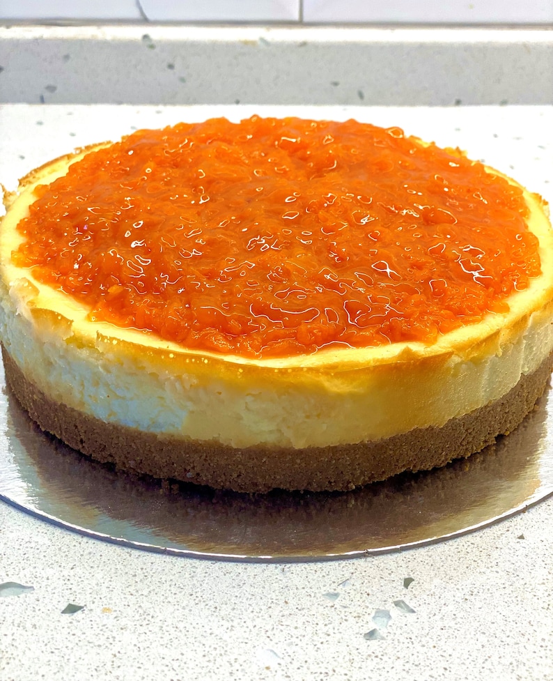 May include: A round cheesecake with a layer of orange marmalade on top. The cheesecake is on a silver plate.