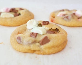 May include: Three chocolate chip cookies with marshmallows and chocolate chips on top. The cookies are on a white surface.