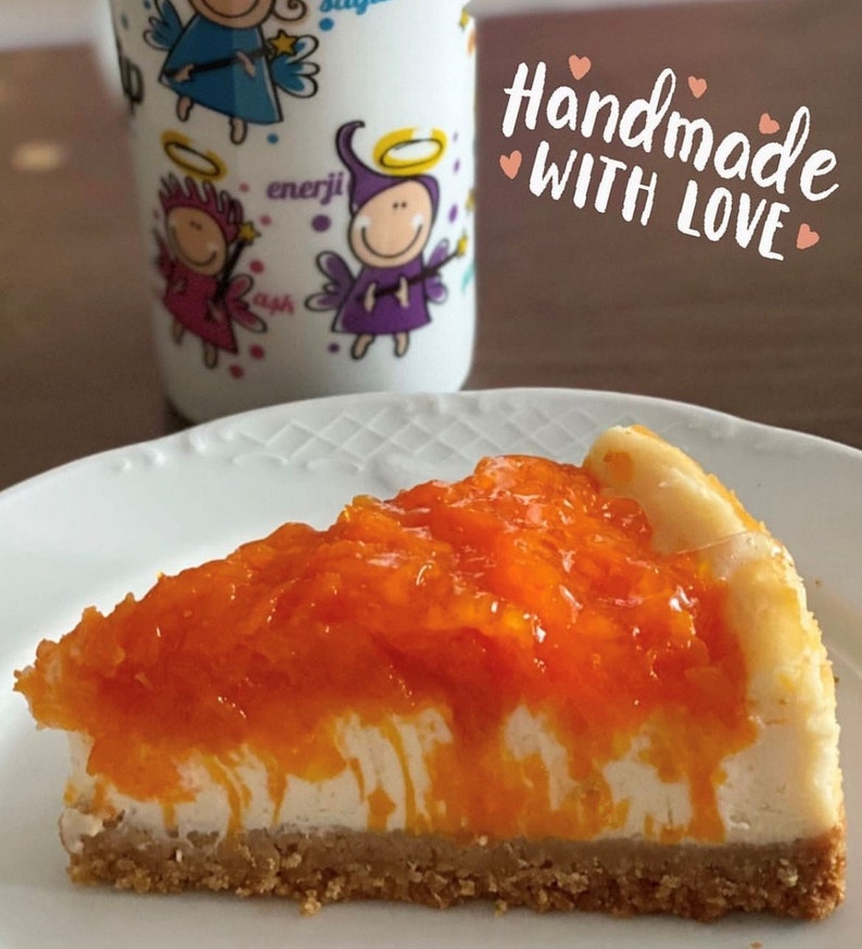 May include: A slice of cheesecake topped with a layer of orange marmalade. The cheesecake has a creamy white filling and a graham cracker crust. The text "Handmade with Love" is written in pink on a brown background.