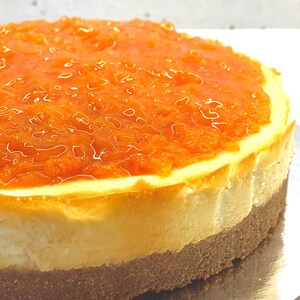 May include: A round cheesecake with a layer of orange marmalade on top. The cheesecake is on a silver plate.