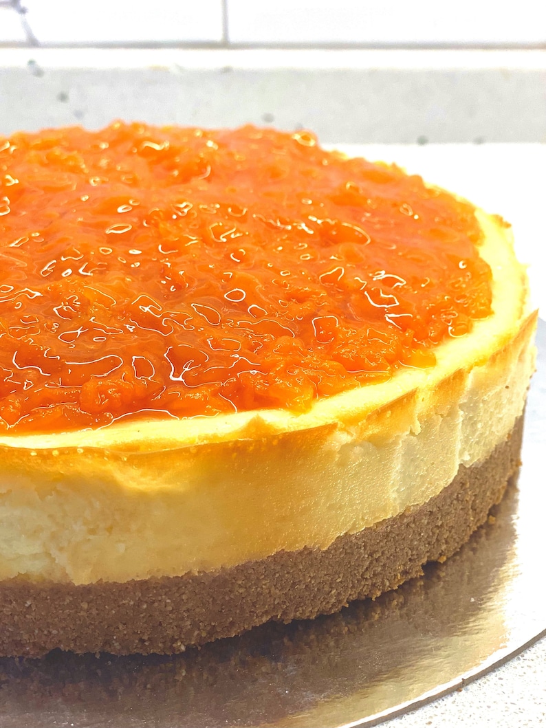 May include: A round cheesecake with a layer of orange marmalade on top. The cheesecake is on a silver plate.