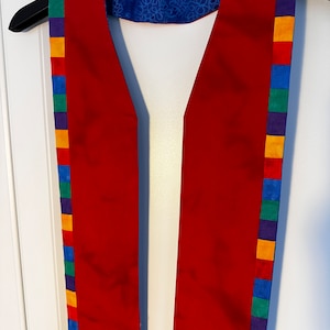 May include: A red stole with a rainbow trim. The trim is made of colorful squares. The stole is hanging on a hanger.