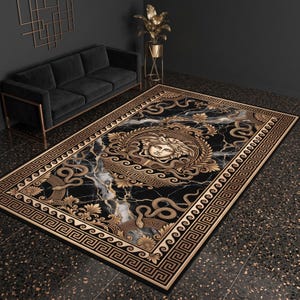 Könnte beinhalten: Ein rechteckiger Teppich mit schwarzem Marmormuster und goldfarbenen Akzenten. Das Design umfasst ein zentrales kreisförmiges Medaillon mit einem von Medusa inspirierten Gesicht, umgeben von verschlungenen Ornamenten, Schlangenmotiven und einer Mäanderbordüre.
