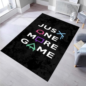 Può includere: Tappeto nero e bianco con il testo "JUST ONE MORE GAME" in bianco. Il testo è circondato da simboli colorati di controller per videogiochi.