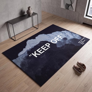 Può includere: Un tappeto rettangolare con una grafica di cielo nuvoloso scuro in tonalità di nero e grigio. Il testo "KEEP OFF" è stampato in grandi lettere maiuscole bianche al centro. Un testo aggiuntivo nell'angolo recita "IKEA c/o Virgil Abloh Flooring Älmhult, Sweden © 2019".