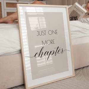 May include: A framed print with a beige background and black text that reads "Just one more chapter". The print is leaning against a white bed with a person lying on it.