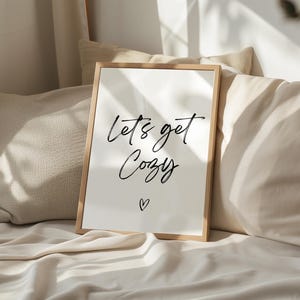 Puede incluir: Un estampado enmarcado con las palabras "Let's get Cozy" en escritura negra, con un pequeño corazón debajo. El estampado tiene un marco de madera clara y está apoyado contra almohadas y ropa de cama en tonos neutros. La imagen evoca una sensación de comodidad y relajación.
