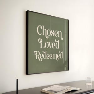 May include: A framed print with the words "Chosen, Loved, Redeemed" in a decorative font. The print is olive green with a black frame, hanging above a black console table. A candle, a book, and a glass are on the table.