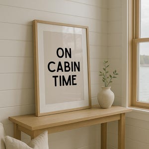 May include: A framed print with the text "ON CABIN TIME" in black. It's displayed on a light wood console table, next to a small vase with greenery. The room has white shiplap walls and a window with a view.
