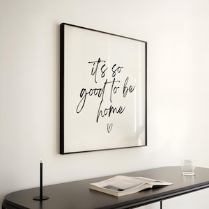Op de afbeelding: Zwarte ingelijste wandkunst met de tekst "It's so good to be home" in witte cursieve letters met een klein hartje onder de tekst.