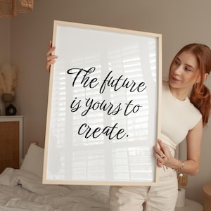 May include: A framed print with the text "The future is yours to create" in elegant black script. The frame is a light wood color, and the print is held by a person. The print is displayed in a well-lit room.