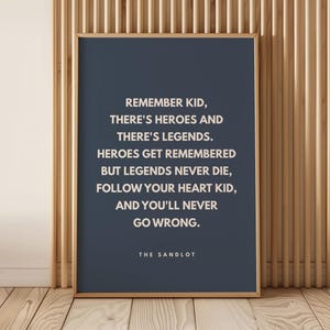 Op de afbeelding: Een rechthoekige wandposter in een dunne houten lijst met een donkerblauwe achtergrond en witte tekst: REMEMBER KID, THERE'S HEROES AND THERE'S LEGENDS. HEROES GET REMEMBERED BUT LEGENDS NEVER DIE, FOLLOW YOUR HEART KID, AND YOU'LL NEVER GO WRONG. THE SANDLOT.