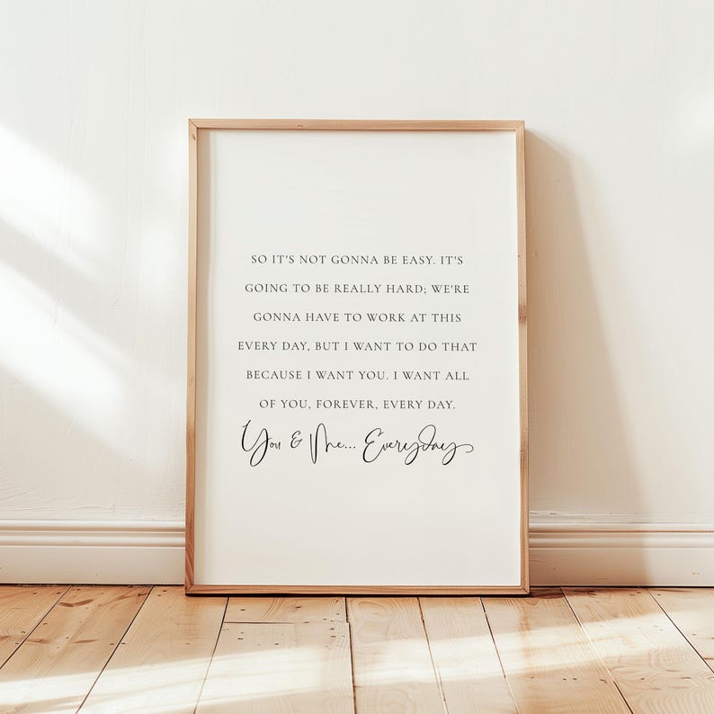 May include: A framed print with a white background and black text. The text reads, "So it's not gonna be easy... You & Me... Everyday." The frame is a light brown color, and the print is displayed on a wooden floor.