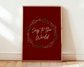 Può includere: Una stampa incorniciata con sfondo rosso scuro. La stampa presenta una ghirlanda circolare di foglie verdi e piccole bacche bianche. Le parole "Joy to the World" sono scritte in elegante corsivo all'interno della ghirlanda. La cornice è di colore marrone chiaro.