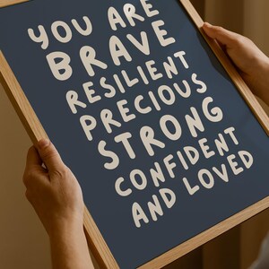 May include: A framed print with a navy blue background and white text. The text reads "YOU ARE BRAVE RESILIENT PRECIOUS STRONG CONFIDENT AND LOVED". The frame is made of light-colored wood.