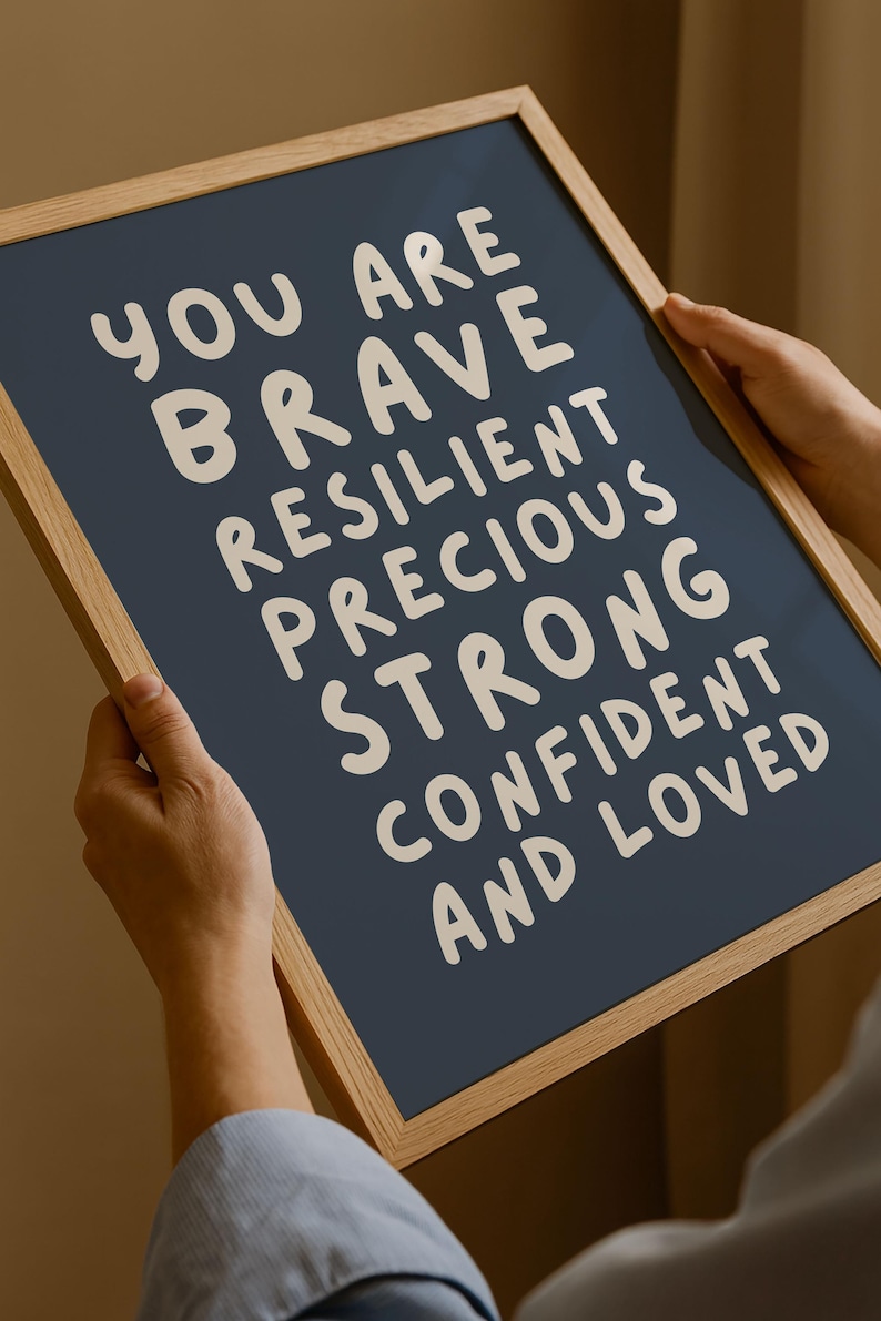 May include: A framed print with a navy blue background and white text. The text reads "YOU ARE BRAVE RESILIENT PRECIOUS STRONG CONFIDENT AND LOVED". The frame is made of light-colored wood.