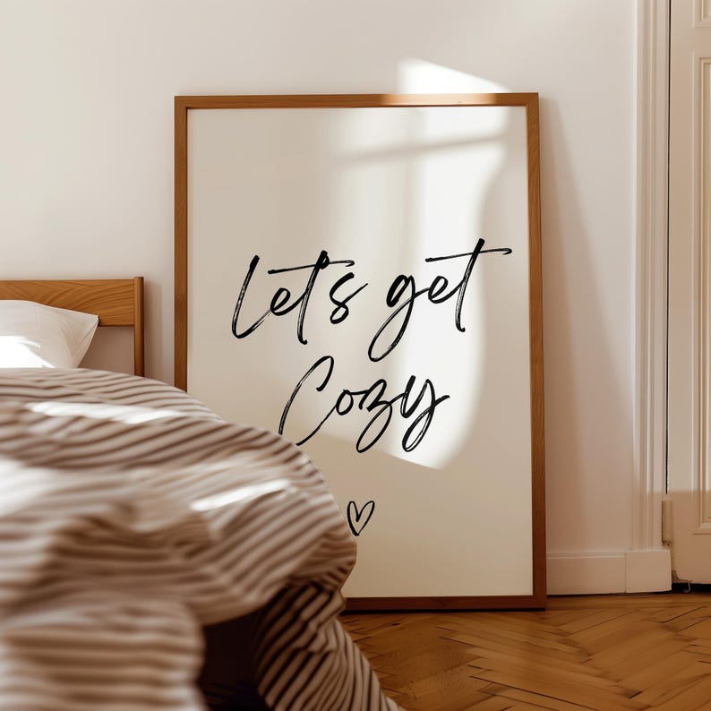 May include: A framed print with the words "Let's get Cozy" in black script, with a small heart below. The print has a cream-colored background and a wooden frame. The image is in a bedroom setting, with a bed and striped bedding visible.