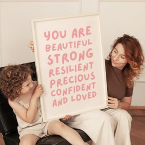 May include: A white framed poster with pink text that reads "You are beautiful strong resilient precious confident and loved". A woman and a child are holding the poster.