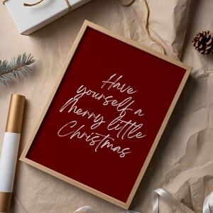 May include: A framed print with a deep red background and the words "Have yourself a Merry little Christmas" in white script. The frame is a light wood color. The image is on a neutral-toned surface with a white gift box and ribbon.