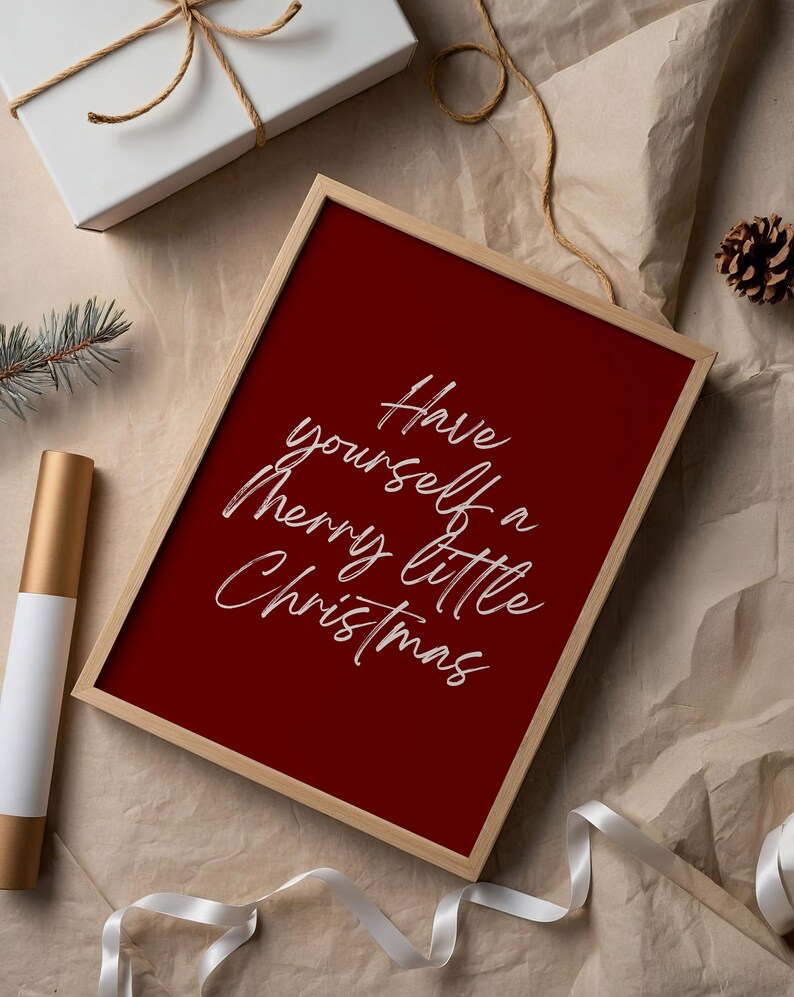 May include: A framed print with a deep red background and the words "Have yourself a Merry little Christmas" in white script. The frame is a light wood color. The image is on a neutral-toned surface with a white gift box and ribbon.