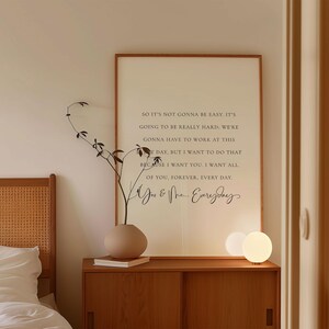 May include: A framed print with a quote in black text: "SO IT'S NOT GONNA BE EASY... I WANT ALL OF YOU, FOREVER, EVERY DAY." The print is displayed above a wooden cabinet with a vase and a small lamp.