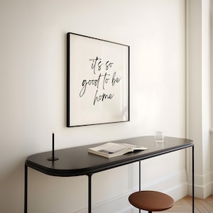 May include: A black framed print with the text "it's so good to be home" hanging on a white wall above a black table with a book and a glass on it.