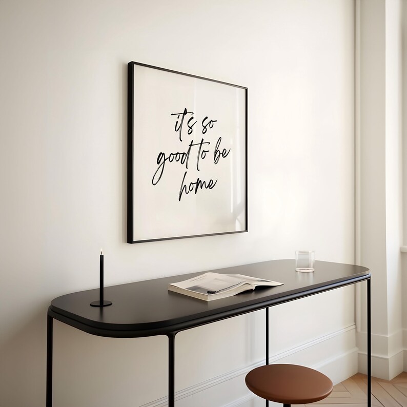May include: A black framed print with the text "it's so good to be home" hanging on a white wall above a black table with a book and a glass on it.