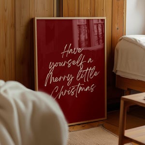 May include: A framed print with a deep red background and the white cursive text "Have yourself a Merry little Christmas." The frame is a light wood color. The print is leaning against a wooden wall.